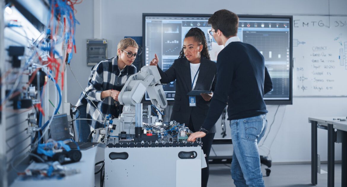 In Robotics Development Laboratory: Black Female Teacher and Two Students Work With Prototype of Robotic Hand. Young Student Telling Something with Smile and Brainstorming with Her Team.