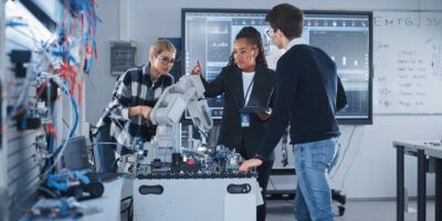 In Robotics Development Laboratory: Black Female Teacher and Two Students Work With Prototype of Robotic Hand. Young Student Telling Something with Smile and Brainstorming with Her Team.