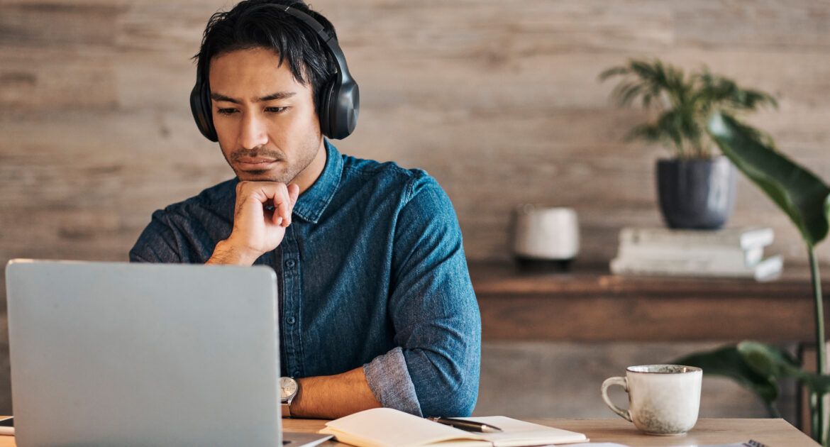 Businessman, headphones and laptop webinar in office with coffee on table, video call or watching video. Zoom call, video conference and male from Canada in online meeting with book and pen on desk.