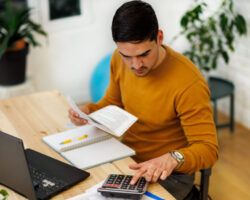 A man working from home using a laptop and calculator to analyze business activities.