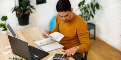 A man working from home using a laptop and calculator to analyze business activities.