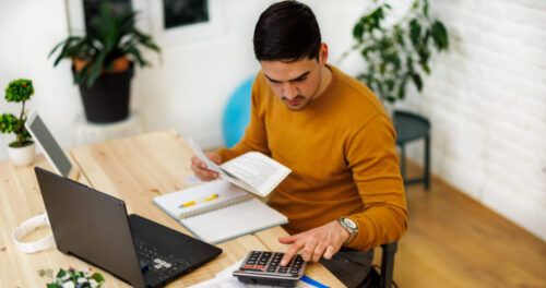 A man working from home using a laptop and calculator to analyze business activities.