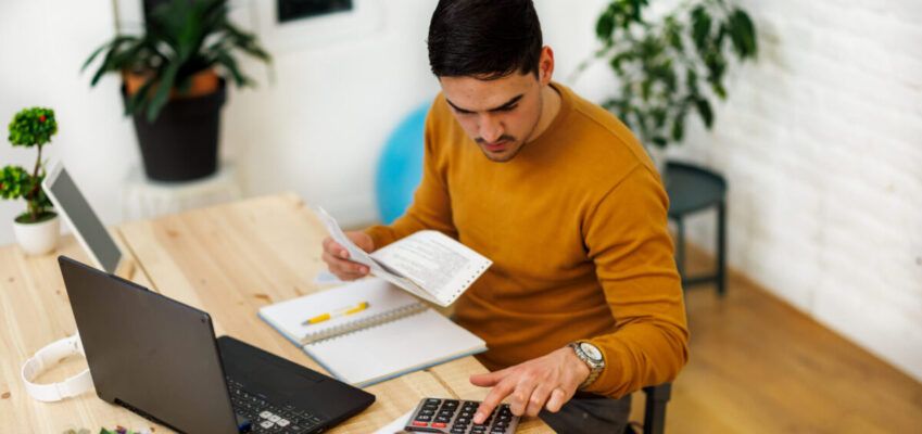 A man working from home using a laptop and calculator to analyze business activities.