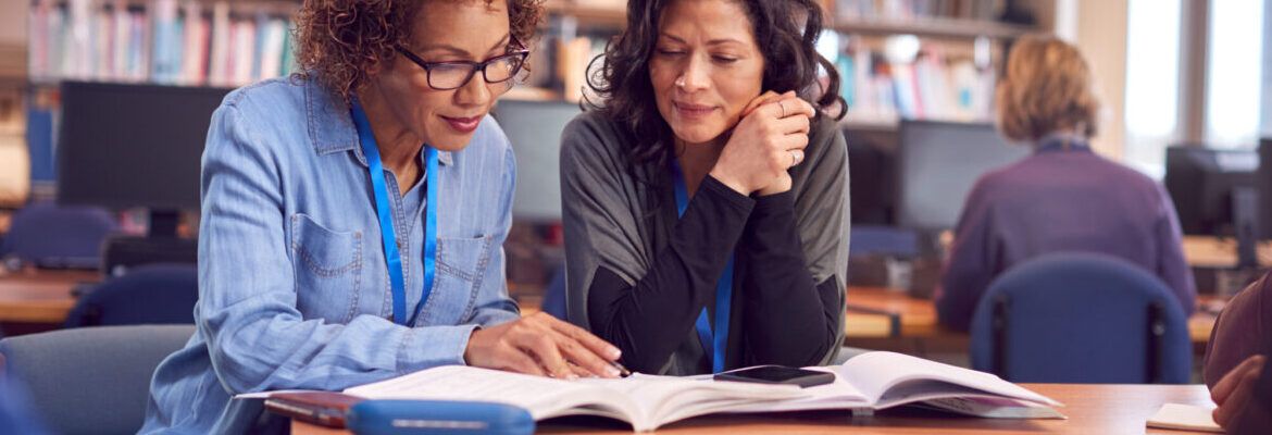 Teacher With Mature Female Adult Student Sitting At Table Working In College Library