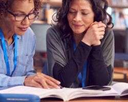 Teacher With Mature Female Adult Student Sitting At Table Working In College Library