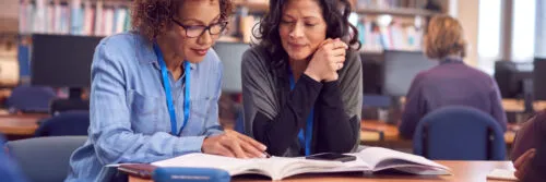 Teacher With Mature Female Adult Student Sitting At Table Working In College Library