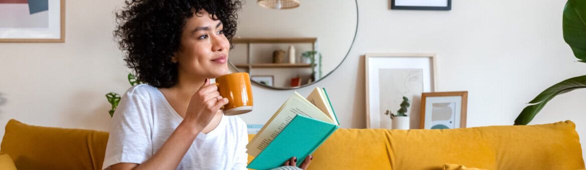 Pensive relaxed woman reading a book at home, drinking coffee sitting on the couch for teacher wellbeing.