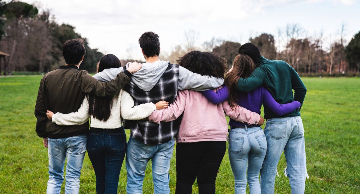 Group of six teenager friends embracing together at the park, rear view