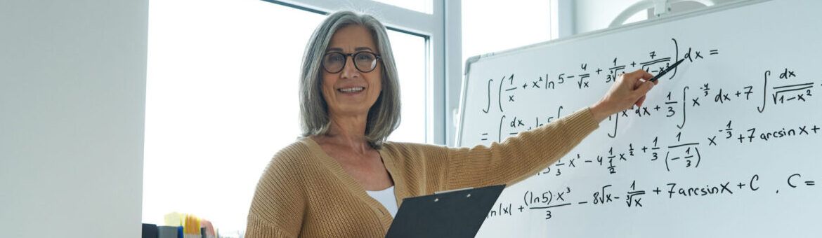 Cheerful senior woman teaching maths while pointing whiteboard at classroom