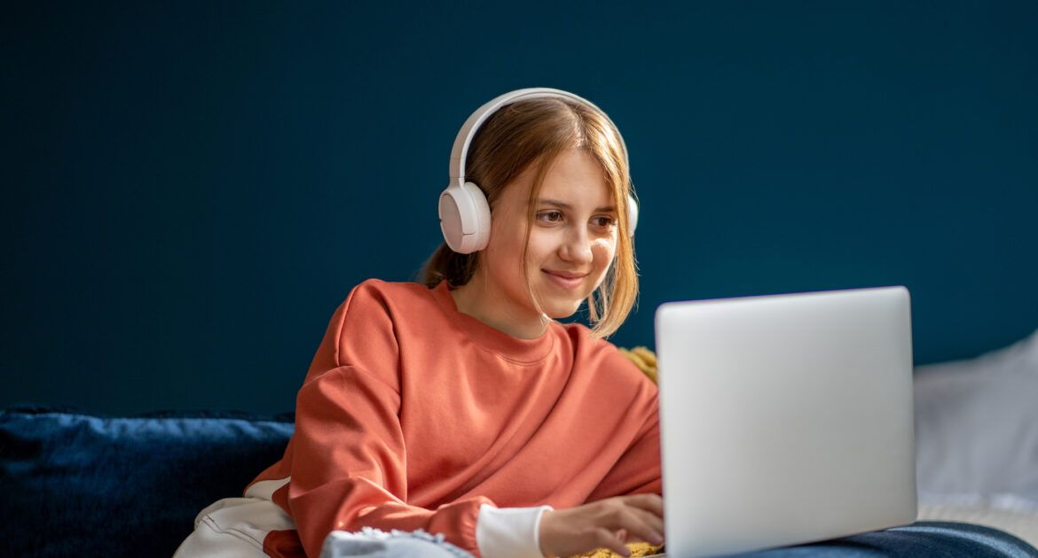 Smiling teenage girl wearing headphones typing on laptop engaging in online teaching