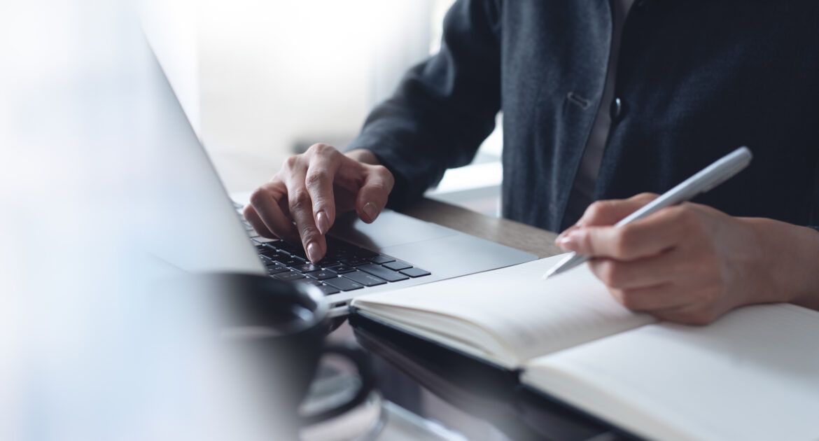 Business woman hands with a pen writing on notebook planner and searching laptop for teaching jobs