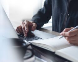 Business woman hands with a pen writing on notebook planner and searching laptop for teaching jobs