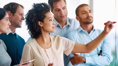 Business people, writing and coaching with tablet for planning, tasks or agenda on glass board at office. Group of young and creative employees taking notes for discussion, brainstorming or startup