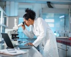 Female Scientist Looking Under Microscope And Using Laptop In A Laboratory