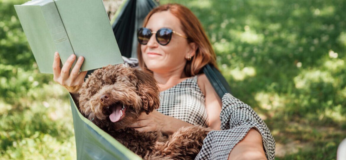 Woman reading book relaxing in hammock with her fluffy brown dog on sunny day.