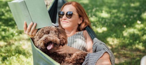 Woman reading book relaxing in hammock with her fluffy brown dog on sunny day.