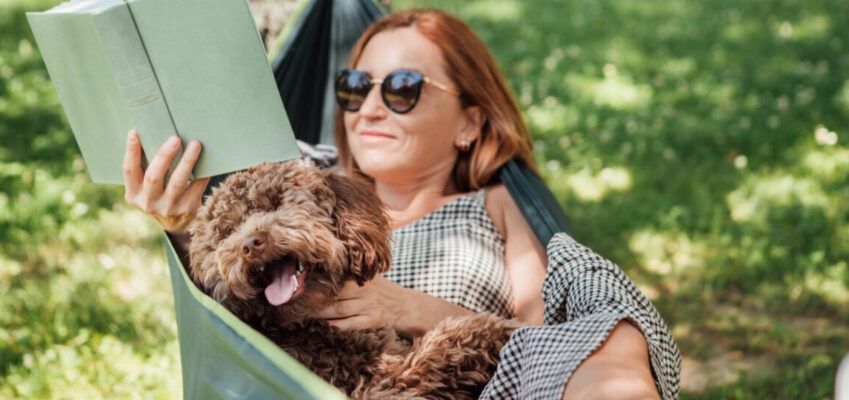 Woman reading book relaxing in hammock with her fluffy brown dog on sunny day.